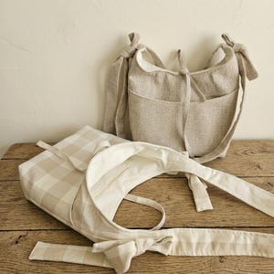 May include: Two beige fabric shoulder bags with knotted straps and pockets. One bag has a checkered pattern, and the other has a solid, textured design. Both bags are displayed on a wooden surface.