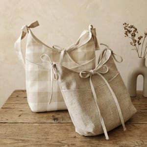 May include: Two beige fabric shoulder bags with bow details. One bag features a plaid pattern, while the other is a solid color. Both bags have a pocket on the front and are displayed on a wooden surface.