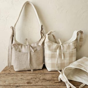 May include: Three beige fabric shoulder bags with pockets and tie closures. One bag is solid, and two have a plaid pattern. The bags are displayed on a wooden surface against a neutral background.