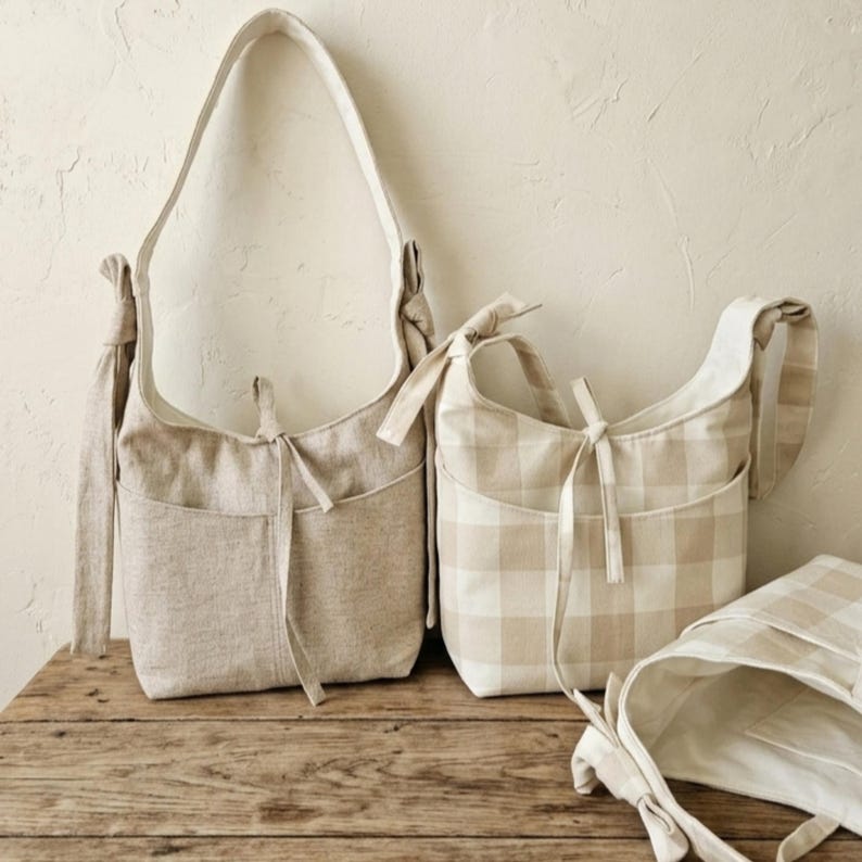 May include: Three beige fabric shoulder bags with pockets and tie closures. One bag is solid, and two have a plaid pattern. The bags are displayed on a wooden surface against a neutral background.