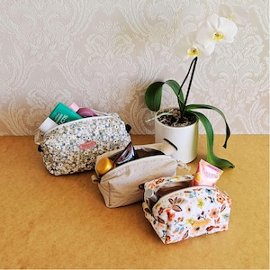 May include: Three cosmetic bags with floral and polka dot patterns are shown. One bag is open, revealing beauty products. A white orchid in a white pot is also in the image. The bags are on a light brown surface.