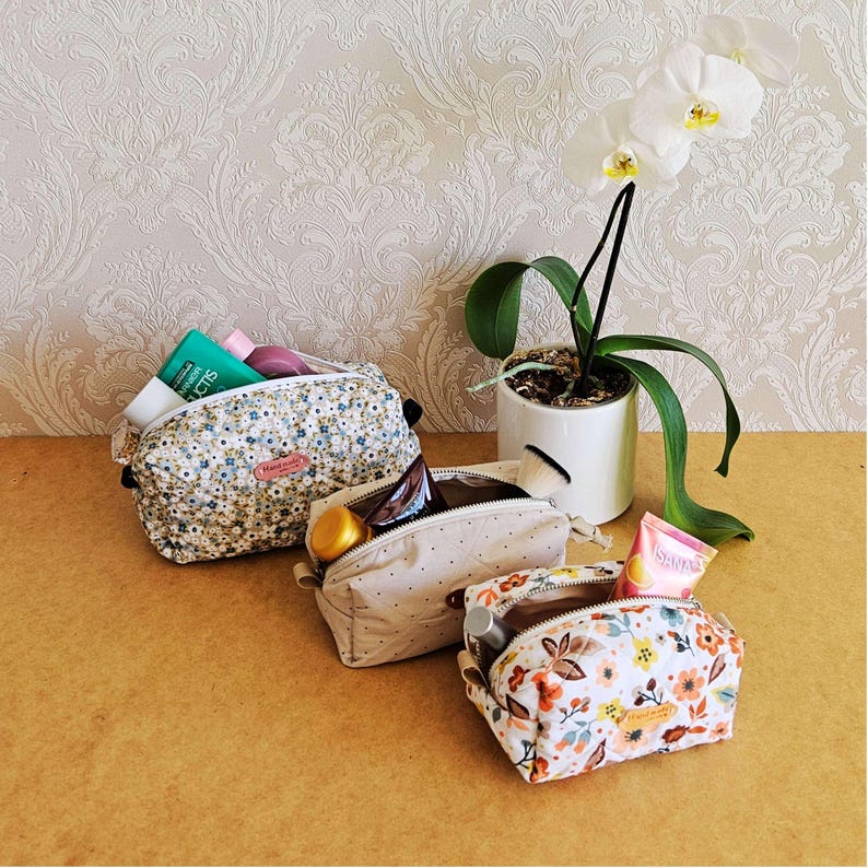 May include: Three cosmetic bags with floral and polka dot patterns are shown. One bag is open, revealing beauty products. A white orchid in a white pot is also in the image. The bags are on a light brown surface.