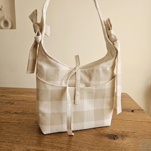 May include: A beige and white checkered shoulder bag with a bow detail on the strap. The bag has a front pocket and a tie closure. The bag is made of a soft fabric and is sitting on a wooden surface.