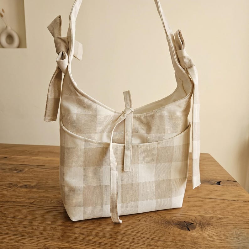 May include: A beige and white checkered shoulder bag with a bow detail on the strap. The bag has a front pocket and a tie closure. The bag is made of a soft fabric and is sitting on a wooden surface.