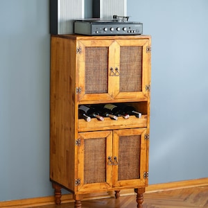 Könnte beinhalten: Ein Holzschränkchen mit zwei Sätzen Doppeltüren, jede mit einer geflochtenen Rattanplatte. Der Schrank hat ein Regal zum Lagern von Weinflaschen und steht auf vier gedrehten Beinen. Ein Vintage-Stereosystem steht auf dem Schrank.
