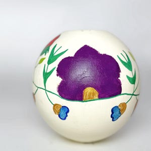 Könnte beinhalten: Handbemaltes, cremefarbenes, kugelförmiges Dekorationsobjekt. Es zeigt eine große lila Blume mit goldenen Akzenten, umgeben von grünen Blättern und Ranken. Blaue und goldene florale Akzente sind ebenfalls vorhanden.