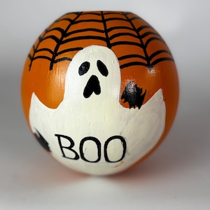 May include: A round, orange Halloween decoration with a hand-painted ghost and spiderweb design. The word "BOO" is written in black letters on the bottom of the ghost. The ghost is white with black eyes and mouth.