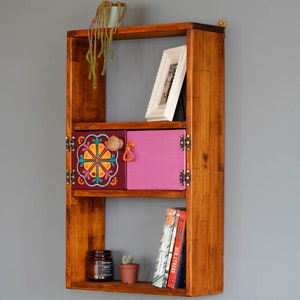 May include: A wooden wall shelf with three shelves. The top shelf has a white picture frame. The middle shelf has two small doors, one with a colorful floral design and the other painted pink. The bottom shelf has a glass jar, a small potted plant, and a stack of books with the title "Real Strand" visible.