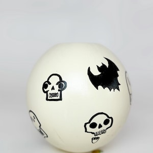May include: A cream-coloured, spherical decorative object with black illustrations. The illustrations include skulls and a bat. The object is likely made of ceramic or wood and is suitable for Halloween or gothic-themed decor.