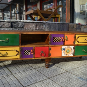 May include: A colorful wooden TV stand with multiple drawers and doors. The drawers are painted in shades of green, yellow, and orange, and the doors are painted in shades of purple, red, and orange. The stand has a unique design with different patterns and textures on each drawer and door.