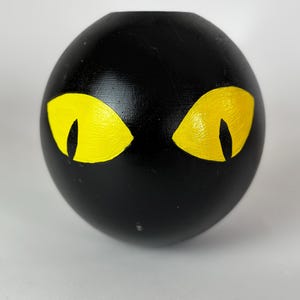 May include: A black, spherical decorative object featuring two yellow, cat-like eyes. The eyes are almond-shaped and painted with a bright, golden colour, contrasting against the dark surface. The object has a smooth, matte finish.