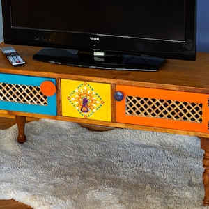 May include: A wooden TV stand with three drawers, painted in bright colors. The drawers have a lattice design and are accented with colorful geometric patterns. The stand has four turned legs.
