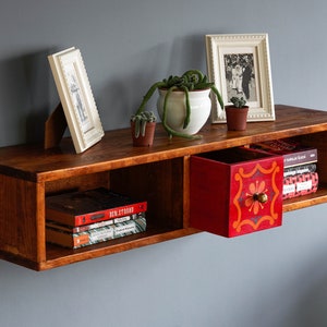 May include: A wooden wall shelf with two open compartments and a red drawer with a floral design. The shelf is decorated with books, plants, and framed photos.
