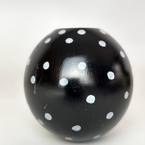 May include: A black, spherical vase with a glossy finish. The vase is decorated with evenly spaced, hand-painted white polka dots. The opening at the top is circular. The vase is a decorative home accent.