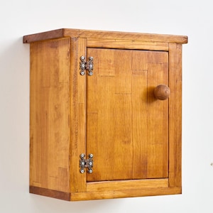 May include: A small wooden cabinet with a single door and a round knob. The cabinet is stained a light brown color and has metal hinges.