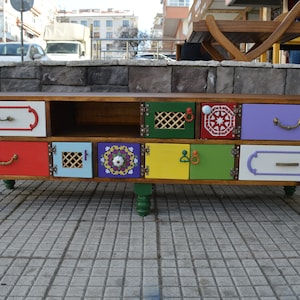 May include: A colorful wooden TV stand with multiple drawers and shelves. The drawers are painted in various colors, including red, green, blue, yellow, and purple. Each drawer has a unique design and a metal handle. The stand has four green wooden legs.
