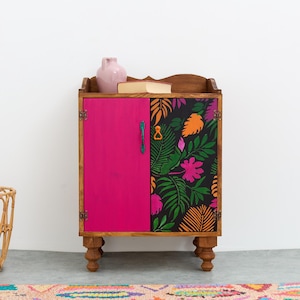 May include: A small wooden cabinet with two doors. The left door is painted a bright pink color. The right door is decorated with a black and white floral pattern. The cabinet has four legs and is sitting on a light gray floor.
