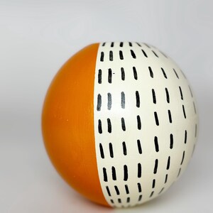 May include: A decorative sphere, half orange and half cream-colored, with a pattern of black dashes. The sphere is a solid color on the orange side and has a hand-painted design on the cream side.