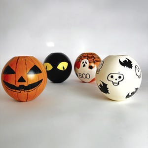 May include: Four spherical Halloween ornaments. One is orange with a carved jack-o'-lantern face. Another is black with yellow cat eyes. The third is orange with a spiderweb and a ghost that says "BOO". The last is cream with bats and a skull.