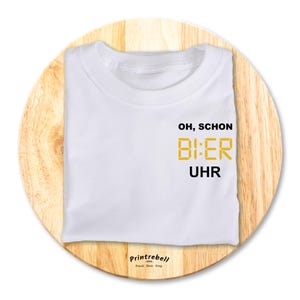 Könnte beinhalten: Weißes T-Shirt mit dem Text "Oh, schon Bier Uhr" in schwarzer und goldener Schrift.