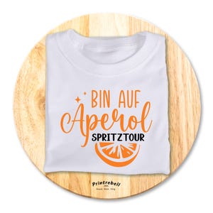 Könnte beinhalten: Weißes T-Shirt mit orangefarbenem Text, der "Bin auf Aperol Spritztour" lautet, und einer Grafik mit einer Orangenscheibe. Der Text "Printrebell" ist unten auf dem Shirt in Schwarz gedruckt.