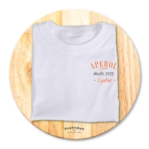 Könnte beinhalten: Weißes T-Shirt mit einem orangefarbenen und grauen Aufdruck, auf dem "Aperol Spritz Malle 2025 - Lydia -" steht.