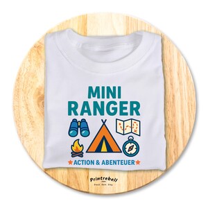 Könnte beinhalten: Weißes T-Shirt mit dem Schriftzug "MINI RANGER" in Türkis, zusammen mit Illustrationen von Fernglas, Zelt, Karte, Kompass und Lagerfeuer. Der Text "ACTION & ABENTEUER" ist ebenfalls enthalten. Das Shirt wird auf einer Holzoberfläche präsentiert.
