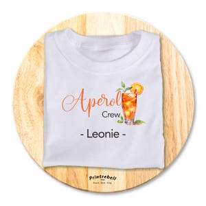 Könnte beinhalten: Weißes T-Shirt mit einer orangenen Aperol Spritz Illustration und dem Text "Aperol Crew - Leonie -" darauf gedruckt.