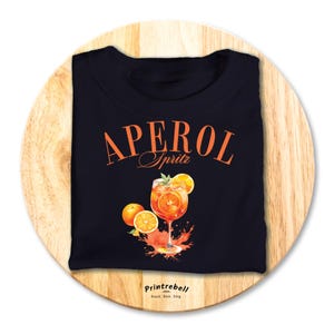 Könnte beinhalten: Schwarzes T-Shirt mit einer orangefarbenen und weißen Illustration eines Aperol Spritz Cocktails. Der Text "Aperol Spritz" ist in Orange auf dem Shirt gedruckt. Der Text "Printrebel" ist in Weiß am unteren Rand des Shirts gedruckt.