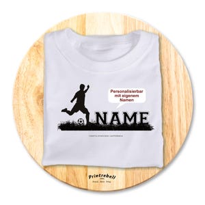 Könnte beinhalten: Weißes T-Shirt mit einer schwarzen Silhouette eines Fußballspielers, der den Ball tritt. Das Wort "NAME" ist schwarz gedruckt, mit einer Sprechblase mit der Aufschrift "Personalizable mit eigenem Namen".