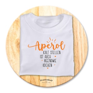 Könnte beinhalten: Weißes T-Shirt mit orangefarbenem Text, der "Aperol Kalt Stellen ist auch irgendwie Kochen" lautet. Der Text ist in einer kursiven Schriftart und hat kleine Herzen drumherum.
