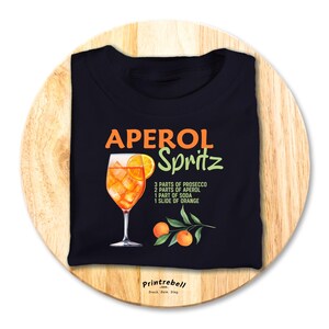 Könnte beinhalten: Schwarzes T-Shirt mit einer Aquarell-Illustration eines Aperol Spritz Cocktails. Der Text "APEROL Spritz" ist in Grün und Orange. Das Rezept für den Cocktail ist unter der Illustration aufgelistet: "3 Teile Prosecco, 2 Teile Aperol, 1 Teil Soda, 1 Scheibe Orange". Der Text "Printrebell" befindet sich unten auf dem Shirt in Weiß.