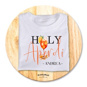 Könnte beinhalten: Weißes T-Shirt mit dem Text "HOLY Aperol" in schwarzer und orangefarbener Schrift. Ein Glas Aperol Spritz ist in Orange und Gelb dargestellt. Der Text "ANDREA" ist in schwarzer Schrift.