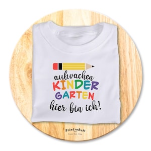 Könnte beinhalten: Weißes T-Shirt mit einer bunten Grafik, auf der "aufwachen kindergarten hier bin ich!" steht und ein gelber Bleistift mit rotem Radiergummi.
