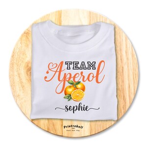 Könnte beinhalten: Weißes T-Shirt mit dem Text "TEAM Aperol sophie" in schwarzer und orangefarbener Schrift. Das Shirt zeigt eine Grafik mit drei Orangenscheiben.