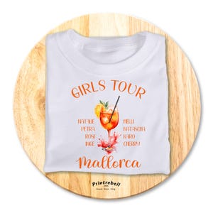 Könnte beinhalten: Weißes T-Shirt mit dem Text "Girls Tour" und einer Aquarell-Illustration eines orangenen Getränks mit Eiswürfeln. Der Text "Mallorca" befindet sich unter der Illustration. Die Namen Natalie, Petra, Rosi, Inge, Melli, Natascha, Karo und Cherry sind auf beiden Seiten der Illustration aufgelistet.