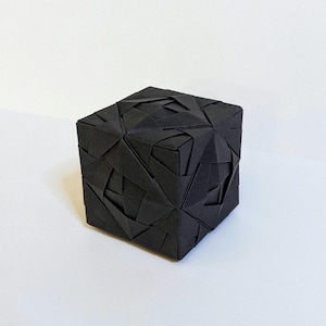 May include: A black origami cube with a complex, woven-like pattern. The geometric form is made of folded paper, creating a textured surface. The cube is displayed on a white background, highlighting its intricate design and form.