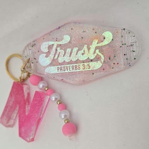 May include: A pink and clear keychain with the word "Trust" and "Proverbs 3:5" printed on it. The keychain has a gold ring and a pink letter "N" charm with a beaded accent.