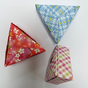 May include: Three origami paper shapes in the image. One is a triangle with a red floral pattern, another is a triangle with a blue and white pattern, and the third is a rectangular shape with a pink and yellow plaid pattern.