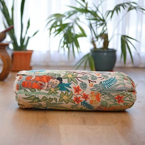 May include: A cylindrical meditation cushion with a floral and patterned design featuring a woman in a red and blue dress. The cushion is on a light wood floor.