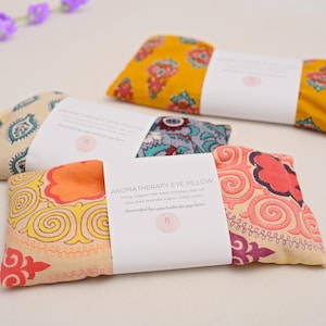 May include: Several aromatherapy eye pillows with colorful floral patterns in yellow, orange, and teal. Each pillow is rectangular and wrapped with a white label. The pillows are filled with organic flax seed and dried lavender. A sprig of purple flowers is in the background.