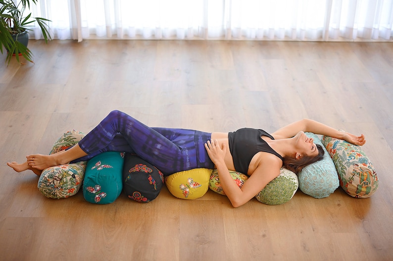 May include: A woman is lying on a row of five colourful cylindrical yoga bolsters. The bolsters are covered in floral and geometric patterns. The woman is wearing purple leggings and a black sports bra.