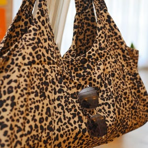 Puede incluir: Un bolso tote grande con un estampado de leopardo en tonos beige y negro. El bolso tiene dos asas y un par de gafas de sol colgando. El bolso está hecho de una tela suave.