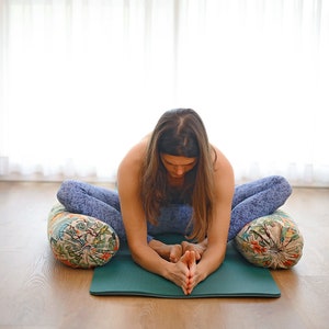 May include: A woman sits in a yoga pose on a green mat with two patterned cushions. She is wearing blue yoga pants and has her hands clasped in front of her.
