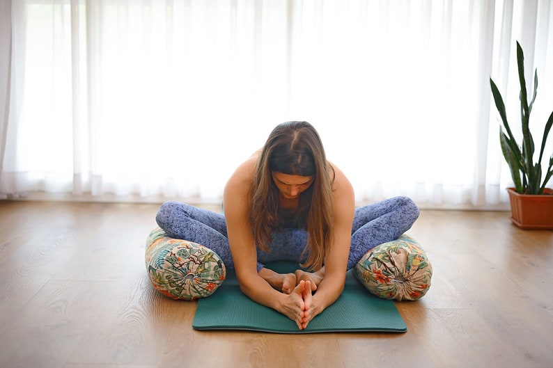 May include: A woman sits in a yoga pose on a green mat with two patterned cushions. She is wearing blue yoga pants and has her hands clasped in front of her.