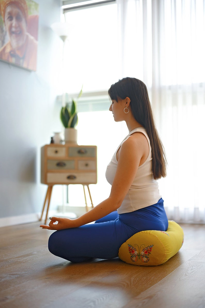 May include: A person sits in a meditative pose on a yellow meditation cushion with a butterfly design. The cushion is on a wooden floor.