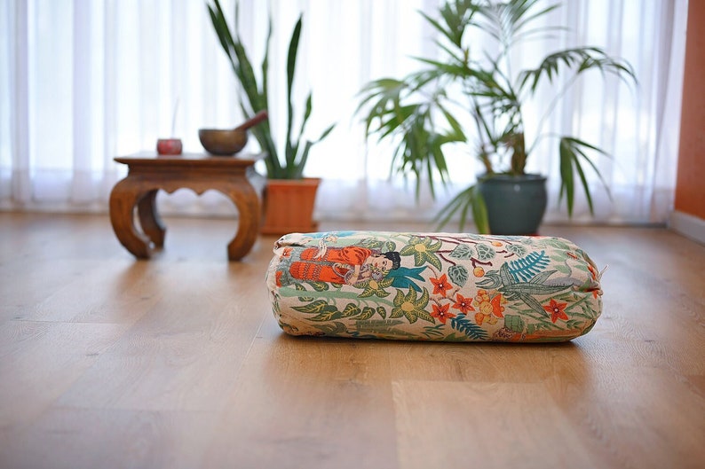 May include: A cylindrical meditation cushion with a floral and patterned design featuring a woman in a red and blue dress. The cushion is on a light wood floor.