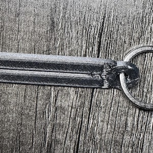 Puede incluir: Un llavero rectangular gris oscuro con un llavero plateado. El llavero tiene una superficie texturizada y una forma rectangular. El llavero es circular y está hecho de metal plateado. El llavero está sobre una superficie de madera.