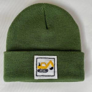 Può includere: Un berretto in maglia color verde oliva a tinta unita con risvolto. Al centro del risvolto si trova una toppa quadrata bianca con bordo nero cucito, che mostra il ricamo di un escavatore da cantiere giallo e grigio.