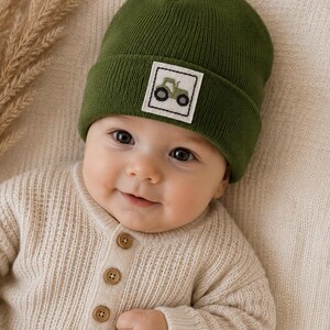Puede incluir: Un gorro de punto verde oliva oscuro con un parche cuadrado blanco que muestra un pequeño tractor verde. El gorro tiene un borde doblado y se combina con una camisa de punto acanalado color crema de manga larga con tres botones marrones.